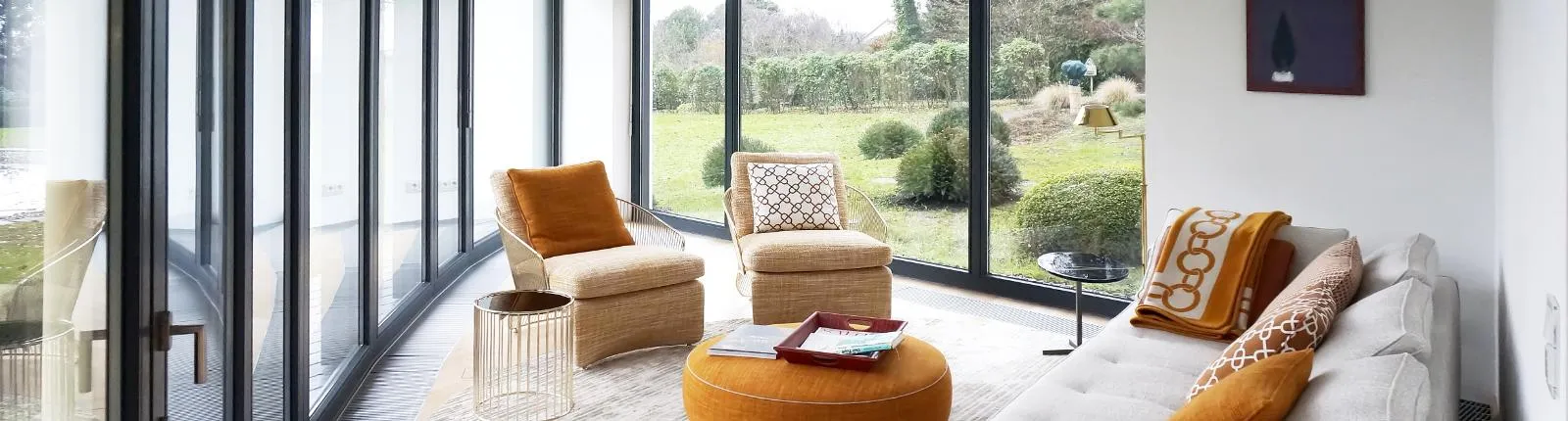 Eleganter Wintergarten mit bodentiefen Fenstern, gemütlicher Sitzgruppe und stilvollen orangefarbenen Akzenten, mit Blick auf einen gepflegten Garten.