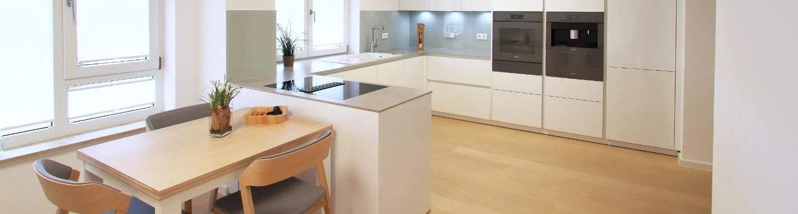 Moderne Wohnküche mit weißen Hochglanzfronten, integrierten Elektrogeräten, Kochinsel und hellem Holzboden, ergänzt durch einen gemütlichen Essbereich.