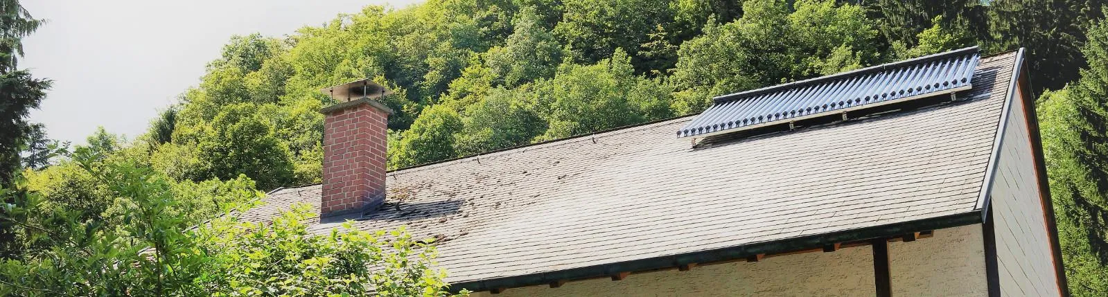 Ein Haus mit Schieferdach in einer waldreichen Umgebung, auf dem Dach sind ein gemauerter Schornstein und eine solarthermische Anlage sichtbar.