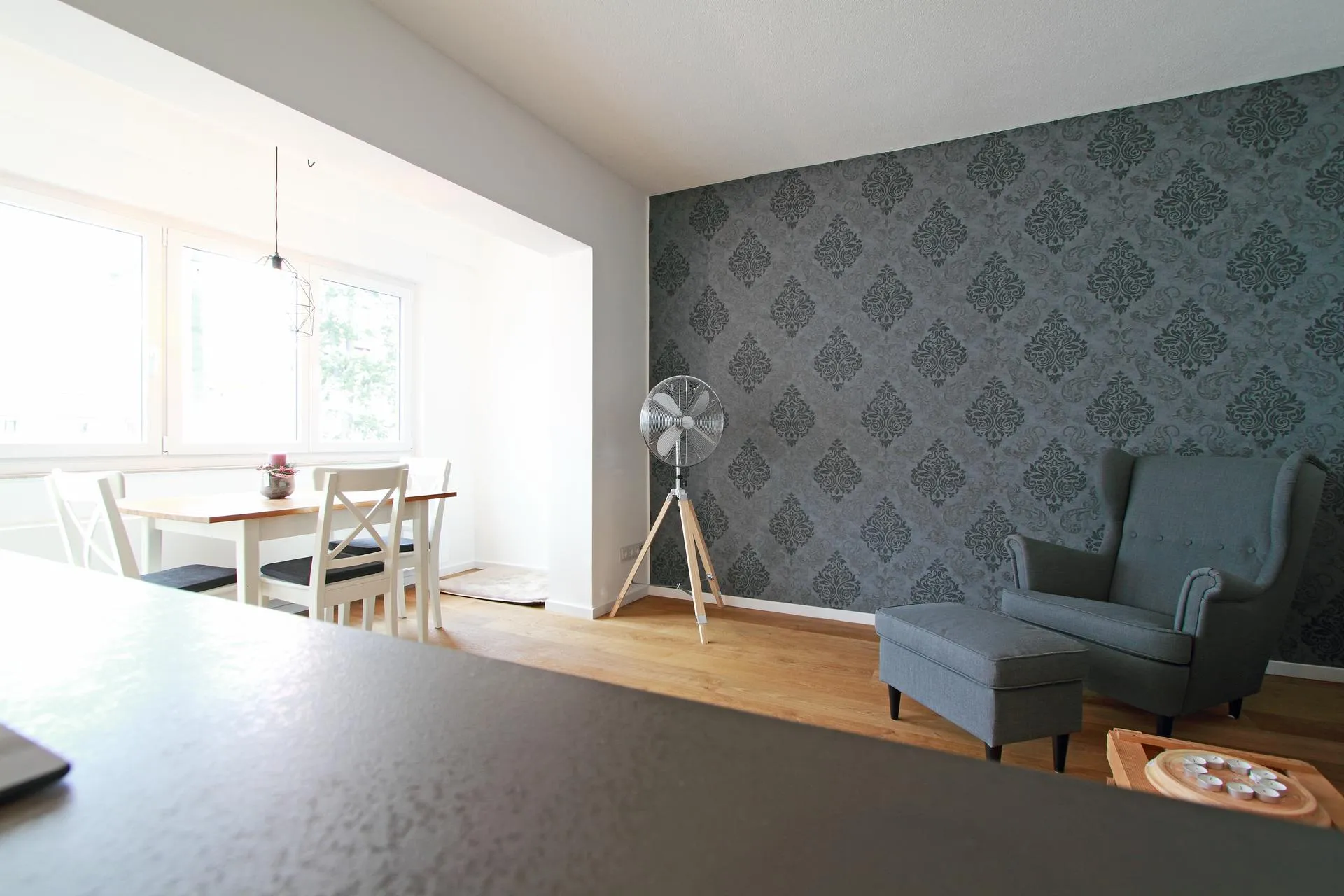 Einladender Ess- und Wohnbereich mit stilvollen Details. Die Wand mit dunkler Tapete im Ornamentstil setzt einen eleganten Akzent, während der gemütliche Sessel mit Hocker zum Entspannen einlädt. Der helle Essplatz mit Holztisch und großen Fenstern schafft eine freundliche, lichtdurchflutete Atmosphäre.