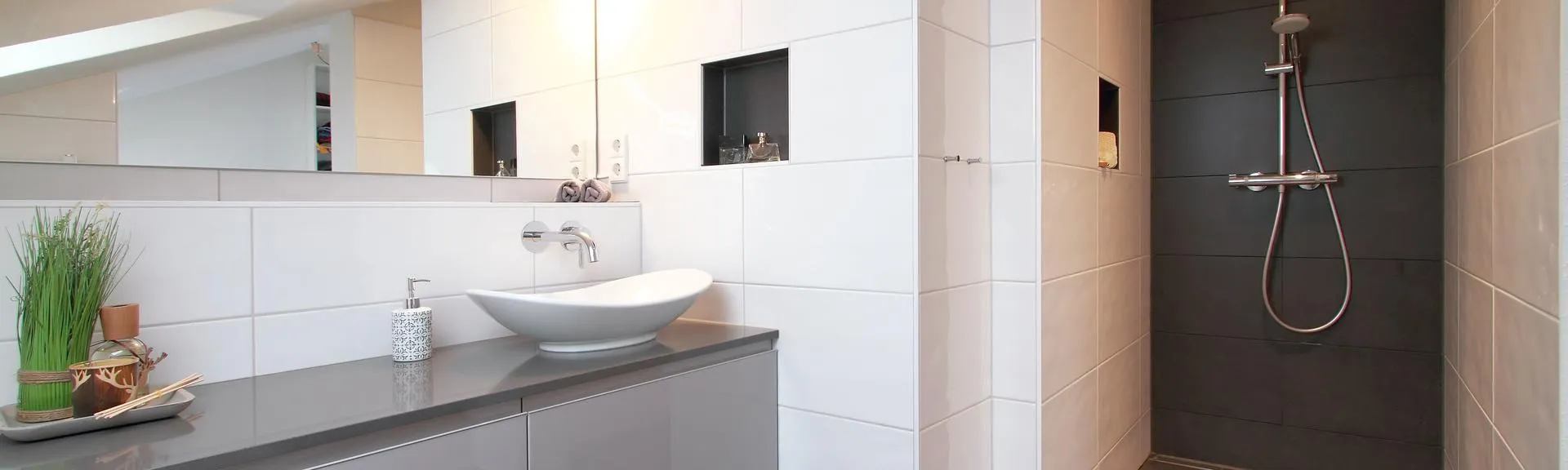 Modernes Badezimmer mit Waschbecken auf grauer Arbeitsplatte, weiß gefliesten Wänden und begehbarer Dusche mit dunklen Fliesen und Regenbrause.