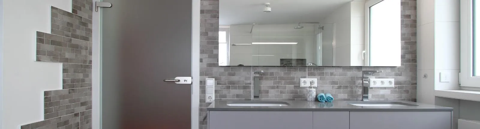 Ein modernes Badezimmer mit einem Doppelwaschbecken und einer grauen Arbeitsfläche, die vor einer Wand aus grauen Mosaikfliesen platziert sind.