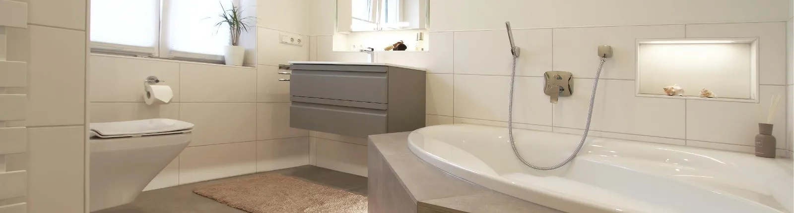Modernes Badezimmer mit hellen Fliesen, einer eingebauten Badewanne mit integrierter Ablage, wandhängendem Waschbecken mit grauen Schubladen, wandhängendem WC, Fenster mit halbtransparentem Sichtschutz und einer Pflanze auf der Fensterbank.