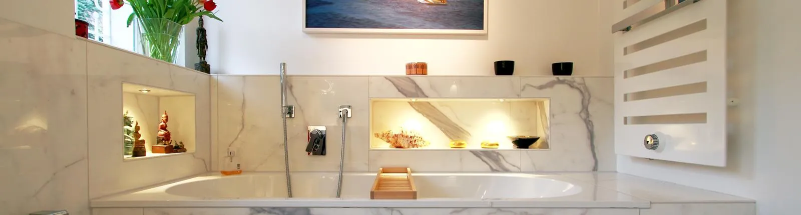 Elegantes Badezimmer mit Marmorausstattung, eingebauter Badewanne, beleuchteten Wandnischen mit Deko-Objekten und einem modernen Heizkörper an der Wand.