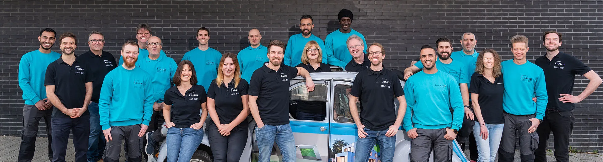 Das komplette Team der Firma Lassen GmbH steht in hellblauen und schwarzen Jacken vor einem Firmen-Fahrzeug und einer dunklen Mauer.