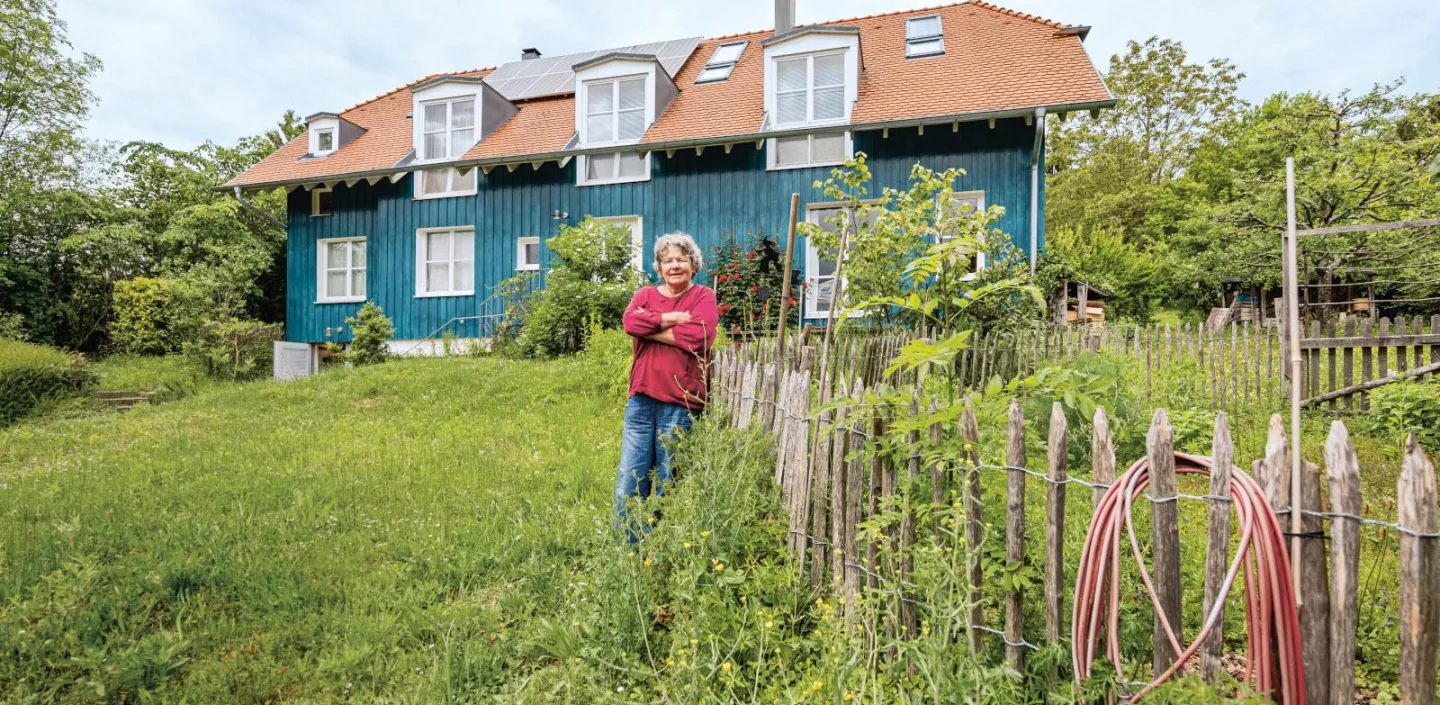 Eine Person steht in einem weitläufigen, grünen Garten vor einem Haus mit blauer Holzfassade und rotem Ziegeldach.