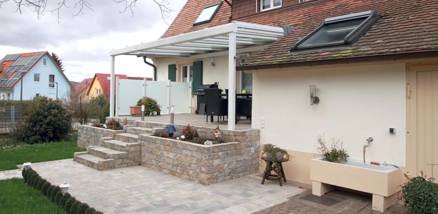 Modern gestaltete Terrasse mit überdachtem Sitzbereich, umgeben von Natursteinmauern und Treppen, eingebettet in einen gepflegten Garten.