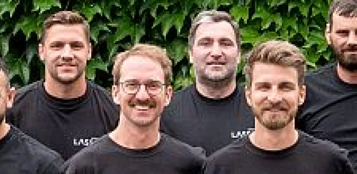 Ein großes Teamfoto der Lassen GmbH zeigt die Mitarbeitenden in schwarzen Firmen-T-Shirts vor einer mit Efeu bewachsenen Wand.