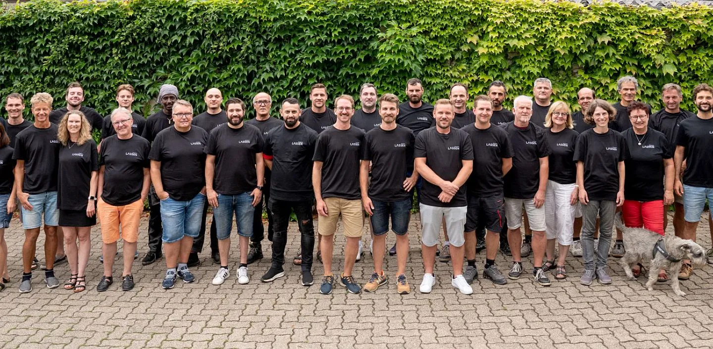 Ein großes Teamfoto der Lassen GmbH zeigt die Mitarbeitenden in schwarzen Firmen-T-Shirts vor einer mit Efeu bewachsenen Wand.