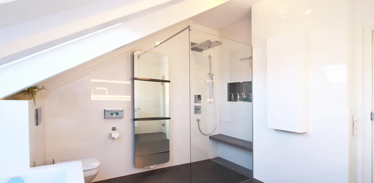 Ein modernes Badezimmer unter einer Dachschräge, mit einer ebenerdigen Dusche, die durch eine Glaswand abgetrennt ist. 