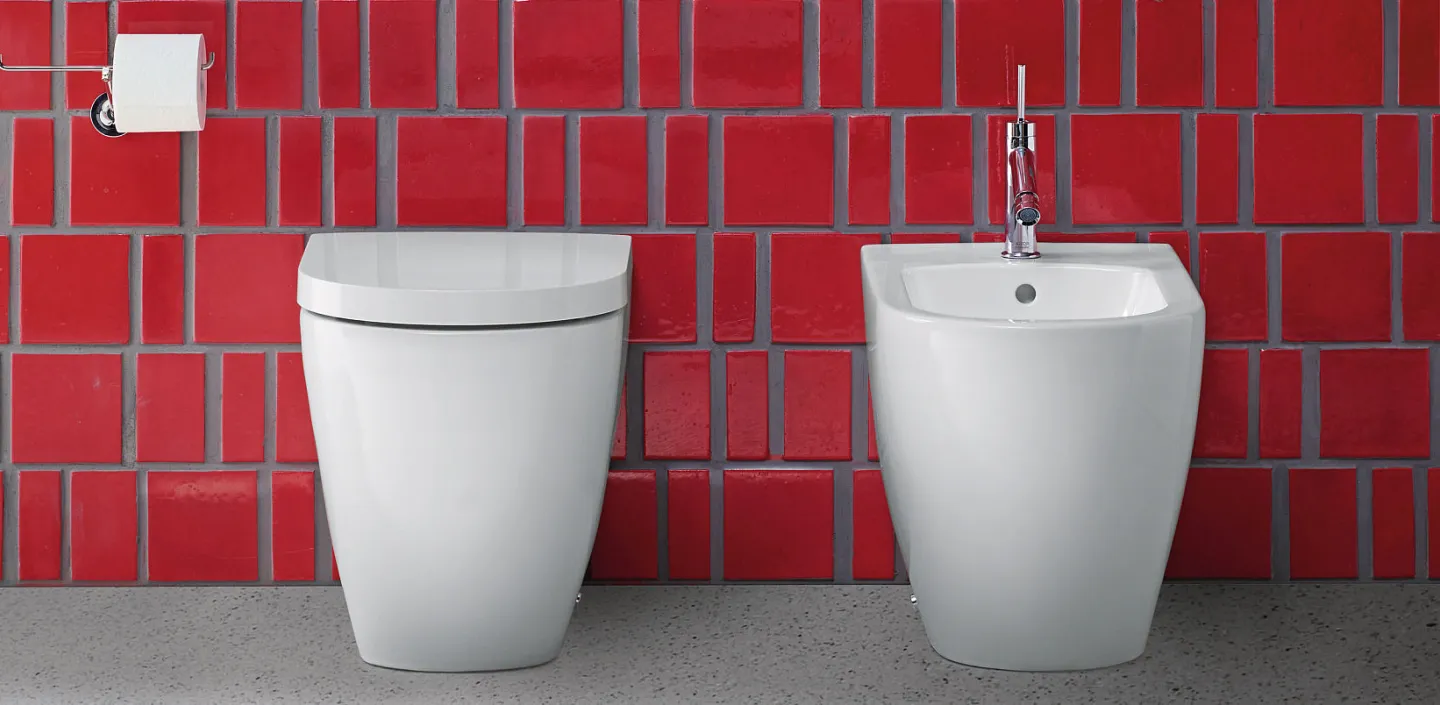 Ein elegantes weißes WC und Bidet, minimalistisch gestaltet, vor einer Wand aus roten Kacheln in kräftigem Glanz.