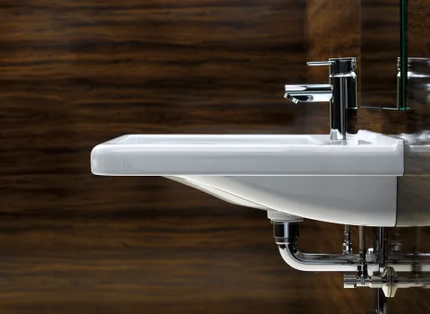 Nahaufnahme eines weißen Waschbeckens mit glänzender Chromarmatur vor einer Wand mit Holzverkleidung. 