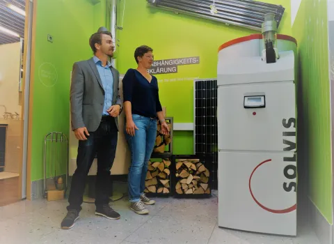 Zwei Personen, ein Mann im Anzug und eine Frau in Freizeitkleidung, stehen vor einer modernen Solvis-Heizung in einem Ausstellungsraum mit grünen Wänden. 