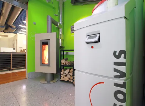 Ein moderner Heizraum mit einer Solvis-Heizung im Vordergrund und einem brennenden Kaminofen links an der grünen Wand.