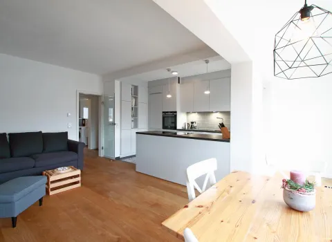 Offener Wohn- und Essbereich mit moderner Einbauküche. Die helle Gestaltung wird durch den warmen Holzfußboden ergänzt. Eine gemütliche Sitzecke, ein Essplatz mit Holztisch und eine stilvolle Beleuchtung sorgen für eine einladende und harmonische Atmosphäre.