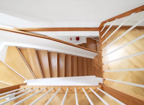 Blick von oben auf eine Wendeltreppe aus hellem Holz mit weißen Geländerstreben. 