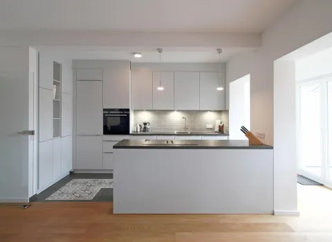 Moderne, offene Küche mit weißen Fronten, grauer Arbeitsplatte und dezenter Beleuchtung, kombiniert mit Holzfußboden und großen Glastüren, die viel Tageslicht hereinlassen.