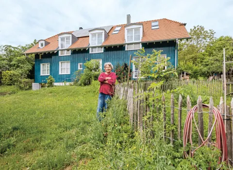 Eine Person steht in einem weitläufigen, grünen Garten vor einem Haus mit blauer Holzfassade und rotem Ziegeldach.