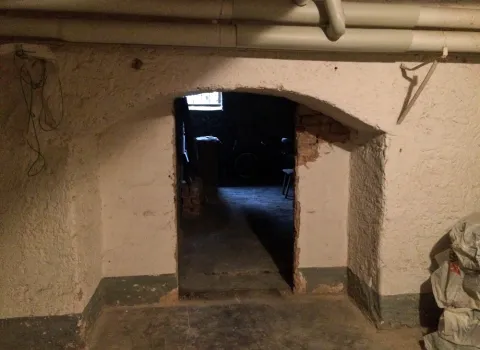 Ein alter Kellerraum mit weiß gestrichenen, rauen Wänden und einem offenen Durchgang, der in einen dunkleren Bereich führt.