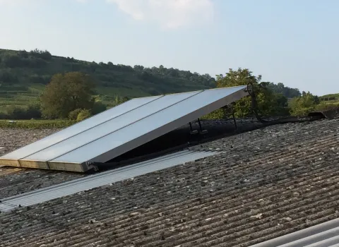 Solaranlage mit schräg montierten Solarmodulen auf einem Wellplattendach in einer ländlichen, hügeligen Umgebung mit Bäumen und grüner Landschaft im Hintergrund unter einem leicht bewölkten Himmel.