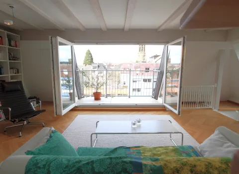 Helles Wohnzimmer mit Zugang zu einer Loggia. Große Glastüren öffnen den Raum zum Balkon, der einen Blick auf die Stadt bietet. 