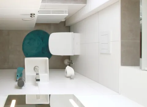 Kompaktes Gäste-WC mit moderner Ausstattung, wandhängendem WC und kleinem Waschbecken, ergänzt durch weiße Fliesen und eine farbliche Akzentuierung mit einem türkisfarbenen Teppich.