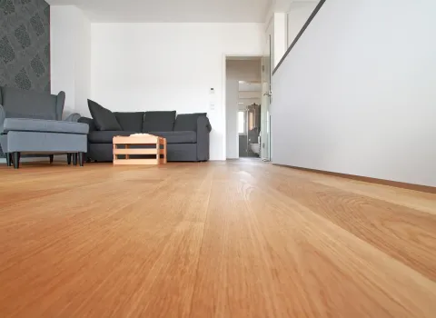 Wohnraum mit hochwertigem Holzboden und moderner Einrichtung. Eine gemütliche graue Sitzgruppe ergänzt den Raum, der durch klare Linien und eine akzentuierte Tapete stilvoll gestaltet ist. Der helle Boden verleiht dem Raum eine warme und einladende Atmosphäre.