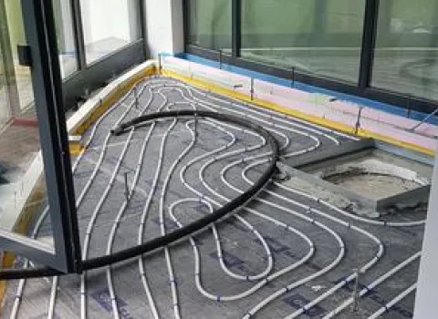 Installationsansicht einer Fußbodenheizung während der Bauphase. Die Heizschlangen sind gleichmäßig auf dem Boden verlegt und mit einer Dämmung sowie Schutzschichten umgeben. Große Fensterfronten sorgen für viel Tageslicht, das den Raum während der Bauarbeiten erhellt.