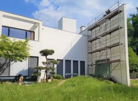 Außenansicht eines modernen Gebäudes während Umbauarbeiten. Eine Seite ist mit einem Baugerüst versehen, während die restliche Fassade in schlichtem Weiß gehalten ist. Der Garten ist von natürlichem Grün geprägt, mit gestutzten Bäumen und einem harmonischen Übergang zwischen Architektur und Natur.