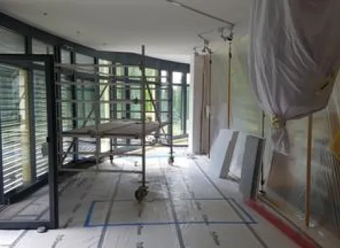 Baustellenansicht eines Innenraums während der Umbauarbeiten. Eine große Fensterfront bietet viel Licht, während Schutzfolien und Gerüste den Arbeitsbereich kennzeichnen. Der Boden ist mit Schutzmaterial abgedeckt, und Werkzeuge sowie Baumaterialien sind vorbereitet, um den Raum zu gestalten.
