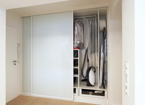 Einbauschrank mit Schiebetüren aus Milchglas, optimal genutzt für die Aufbewahrung von Haushaltsgegenständen wie Staubsauger, Bügelbrett und Kleidung.