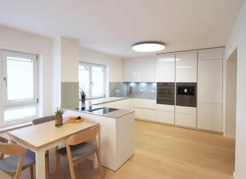 Moderne Wohnküche mit weißen Hochglanzfronten, integrierten Elektrogeräten und einer Kochinsel, kombiniert mit einem Essbereich aus hellem Holz und viel natürlichem Licht.