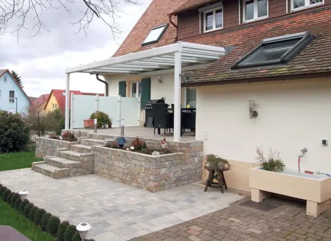 Modern gestaltete Terrasse mit überdachtem Sitzbereich, umgeben von Natursteinmauern und Treppen, eingebettet in einen gepflegten Garten.