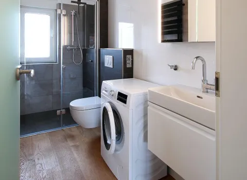 Ein modernes, kompaktes Badezimmer mit einer gläsernen Dusche, einer wandhängenden Toilette und einer Waschmaschine.
