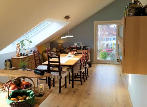 Ein gemütlicher Essbereich unter einer Dachschräge mit einem großen Holztisch und passenden Holzstühlen mit gestreiften Sitzkissen. 