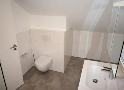 Ein modernes Badezimmer mit einer weißen Wand-Toilette und großformatigen, weißen Wandfliesen. 