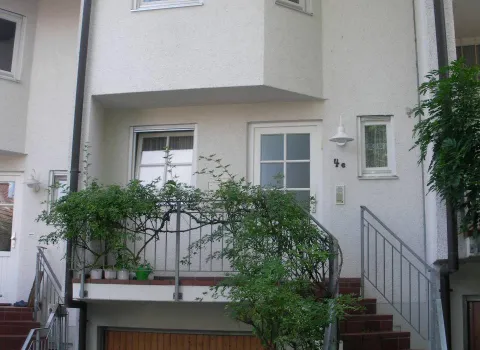 Ein dreistöckiges Reihenhaus mit einer weißen Fassade und einem kleinen Balkon, der von einem Geländer mit Kletterpflanzen umgeben ist.