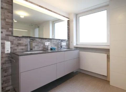 Ein modernes Badezimmer mit einem großen Spiegel, der den Raum optisch vergrößert, und einer grauen Waschtischanlage mit Schubladen.