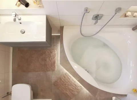 Vogelperspektive eines modernen Badezimmers mit hellen Fliesen, einer gefüllten Eckbadewanne, grauen Badteppichen, einem wandhängenden Waschbecken mit grauem Unterschrank und einem wandhängenden WC. Kleine Details wie ein Deko-Ente und Muscheln in einer Nische runden die Einrichtung ab.