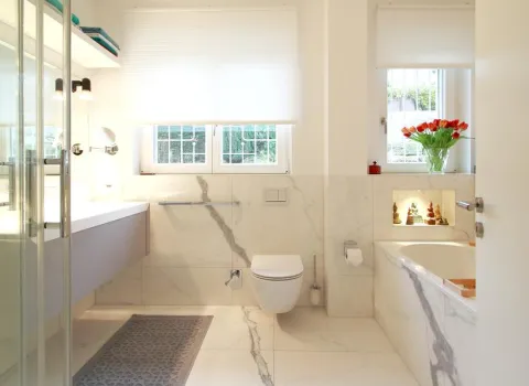 Elegantes Badezimmer in Marmoroptik mit Glasdusche, schwebendem Waschtisch, wandhängender Toilette und Badewanne, ergänzt durch große Fenster mit Blick ins Grüne und dekorative Nischenbeleuchtung.