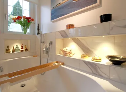 Helles Badezimmer mit Marmordesign, eingebauter Badewanne, dekorativen Wandnischen mit Beleuchtung und Accessoires sowie einem Fenster mit Blumenstrauß im Hintergrund.