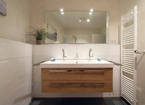 Ein stilvolles Badezimmer mit einem Waschtisch aus Holz, der mit zwei modernen Armaturen und einem großen, rahmenlosen Spiegel ausgestattet ist.