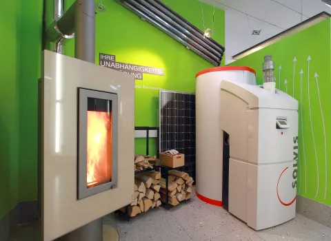 Eine Energieausstellung zeigt einen funktionierenden Pelletofen, Holzvorräte und ein modernes Wärmespeicher-System vor grüner Hintergrundgestaltung.