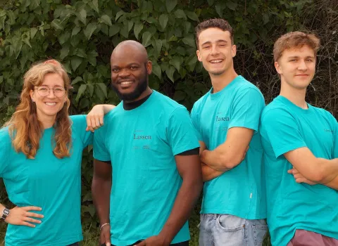 Eine Gruppe von vier jungen Azubis in türkisfarbenen Team-Shirts posiert lächelnd vor einer Hecke.