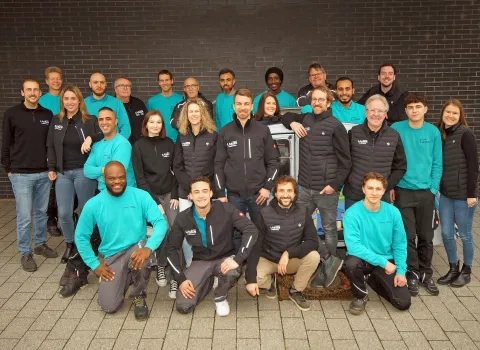 Das komplette Team der Firma Lassen GmbH steht in hellblauen und schwarzen Jacken vor einer dunklen Mauer.