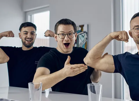 Junge Männer zeigen ihre Muskeln in einem hellen Raum, während ihre Kollegen lachend applaudieren.