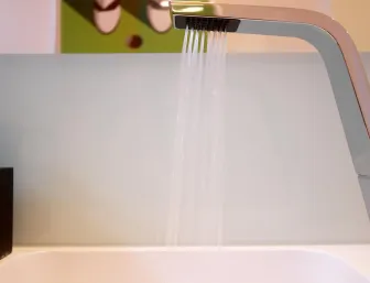 Nahaufnahme eines modernen Wasserhahns, aus dem ein feiner Strahl Wasser in ein Waschbecken fließt. 