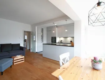 Offener Wohn- und Essbereich mit moderner Einbauküche. Die helle Gestaltung wird durch den warmen Holzfußboden ergänzt. Eine gemütliche Sitzecke, ein Essplatz mit Holztisch und eine stilvolle Beleuchtung sorgen für eine einladende und harmonische Atmosphäre.