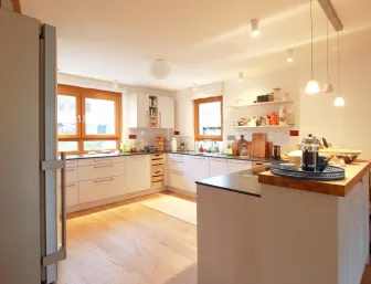Gemütliche Wohnküche mit U-förmiger Arbeitsfläche, weißen Fronten und Holzakzenten, beleuchtet durch warme Pendelleuchten und Tageslicht durch Holzrahmenfenster.