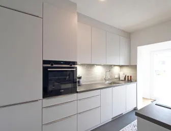 Moderne Einbauküche mit klaren Linien und hochwertigen Materialien. Die matte Oberfläche der weißen Schränke wird durch eine dunkle Arbeitsplatte ergänzt. Dezente Beleuchtung unter den Oberschränken und ein stilvoller Backofen sorgen für eine funktionale und elegante Küchengestaltung.